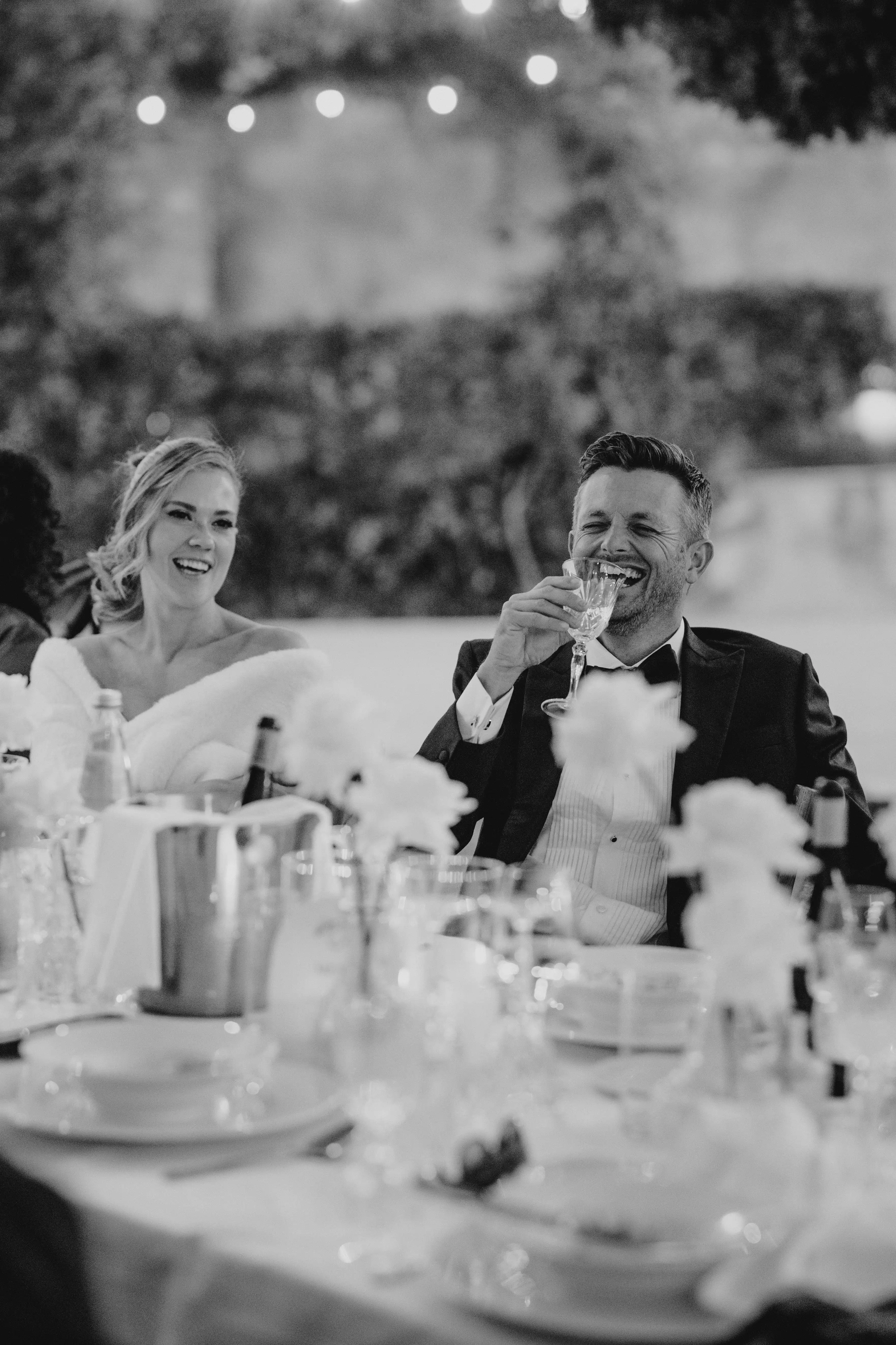 Black and white portrait of bride and groom laughing together during speeches at their wedding dinner reception.